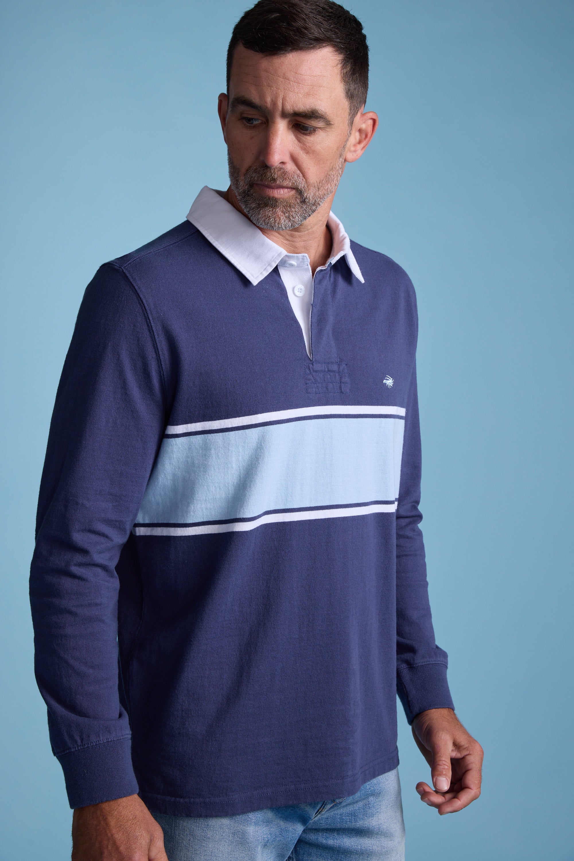 A man with short hair and a beard wears the Barkers Breaker Rugby Jersey, featuring a white collar and horizontal light blue stripe, made from organic cotton, set against a plain light blue background.