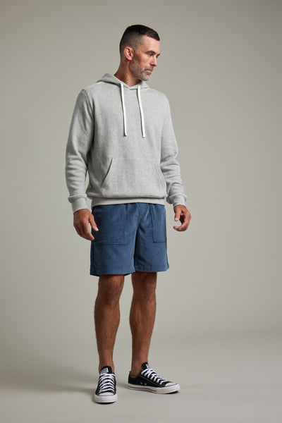 A man stands in a studio wearing the Barkers Inlet Hoody, paired with blue shorts and black low-top sneakers with white soles. He looks to the side with a neutral expression against a plain background.