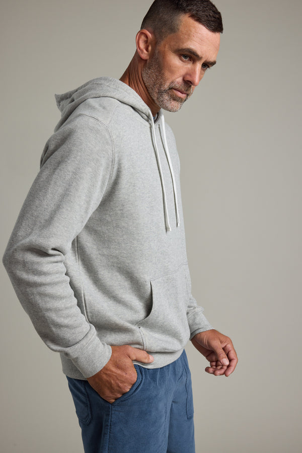 A man with short hair and a beard wears the Barkers Inlet Hoody in light gray and blue pants, standing against a plain, light background with one hand in his pocket, looking down.