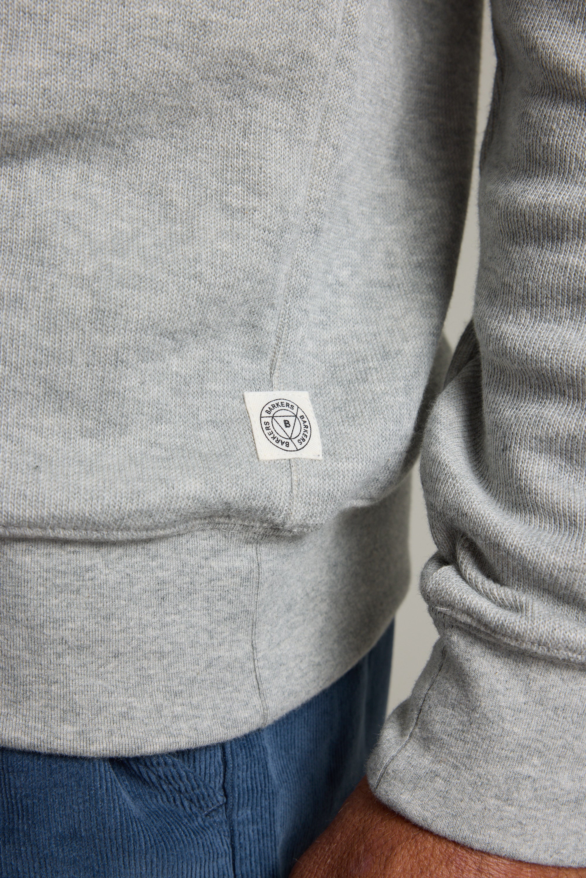 A close-up shows part of a person’s arm and hand wearing the Barkers Inlet Hoody in light grey organic cotton, featuring ribbed sleeves and a small white tag near the hem, paired with blue pants.