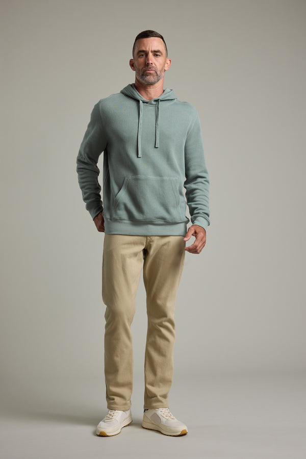 A man stands against a plain background wearing the Barkers Inlet Hoody in light green, paired with beige pants and white sneakers, his hands relaxed by his sides.