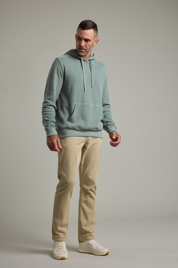 A man wears the Barkers Inlet Hoody in light blue, paired with beige pants and white sneakers, standing against a plain light gray background and gazing slightly to his left with a neutral expression.