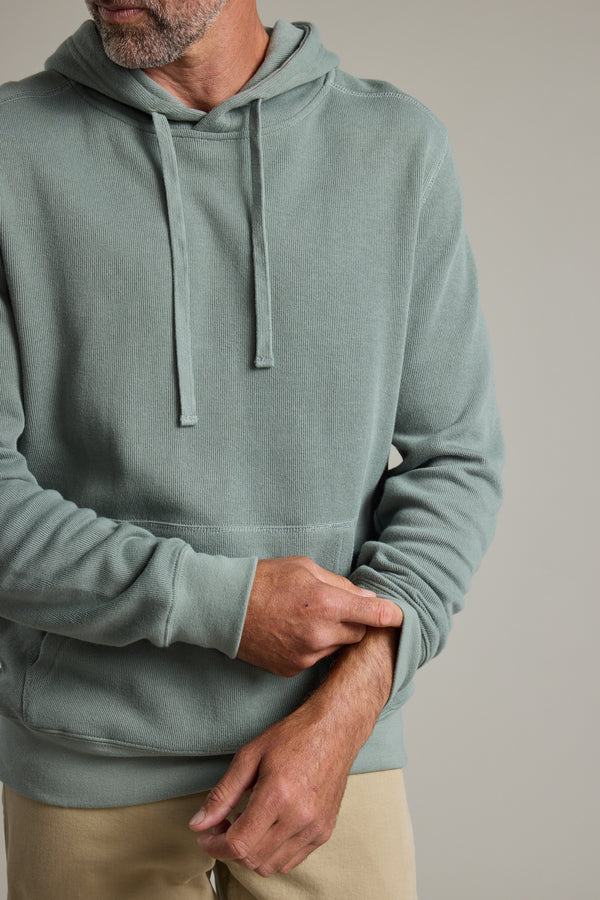 A man wears the Barkers Inlet Hoody in light green with beige pants, rolling up his left sleeve slightly; his face is partially out of frame against a plain light background.