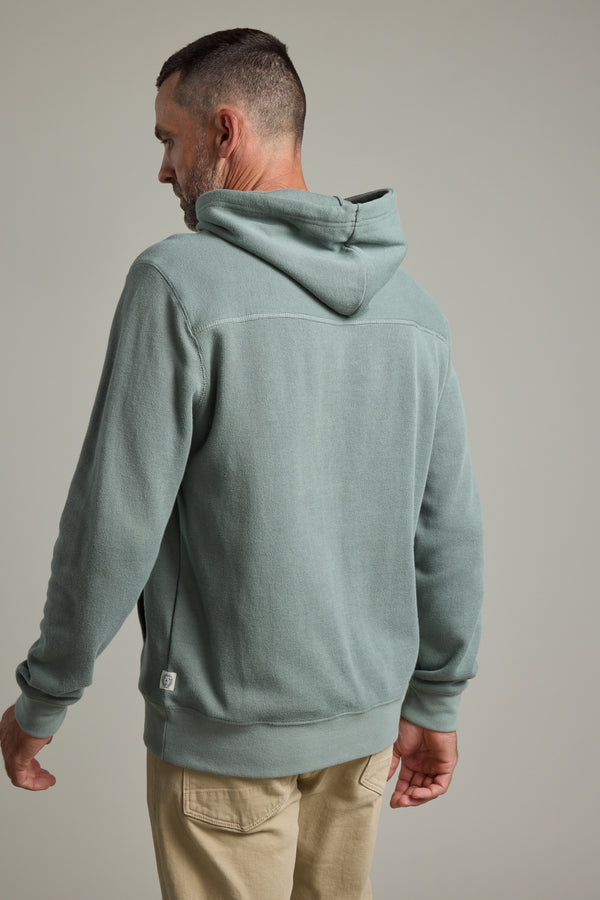 A man faces away from the camera, wearing Barkers’ Inlet Hoody in light green organic cotton and beige pants, set against a plain backdrop.