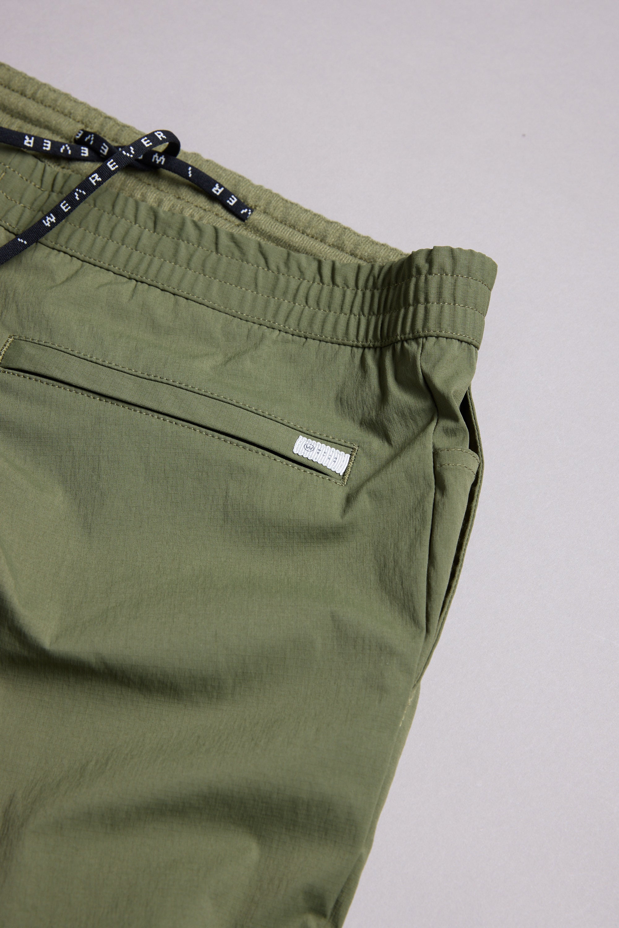 The WearEver Transit Pant in olive green features an elastic waistband, black drawstring with white text, and a zippered pocket. Shown laid flat on a light grey background to showcase the upper portion and pocket detail.
