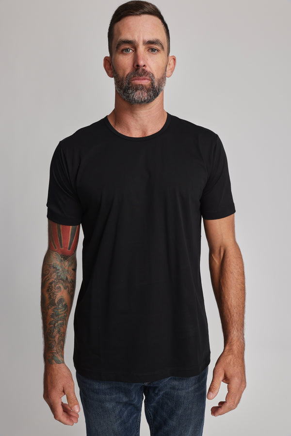 A man with short hair, a beard, and a tattooed left arm stands facing forward in a black Barkers Linton Supima Tee and blue jeans. He has a neutral expression against a plain light background.