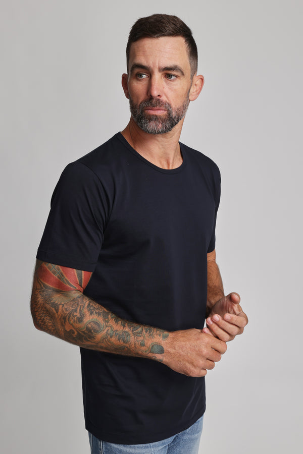 A man with short dark hair, a beard, and tattooed arms wears the Barkers Linton Supima Tee in black with light blue jeans. Standing against a light gray background, he looks left with hands loosely clasped—a true wardrobe staple.