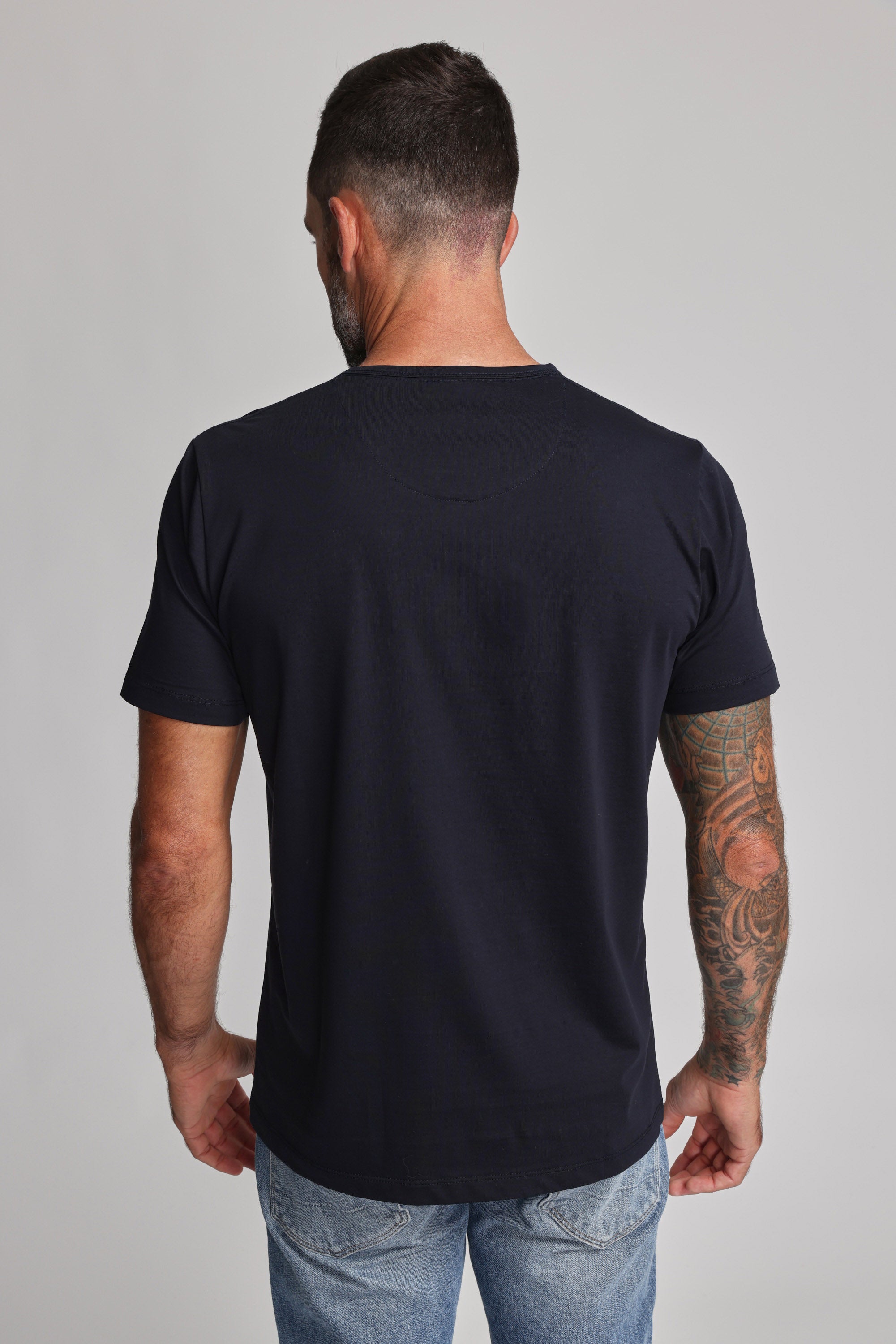 A man stands with his back to the camera, wearing Barkers' Linton Supima Tee in black—crafted from premium mercerised Supima cotton—and light blue jeans. His tattooed left arm, short hair, and beard stand out against a neutral backdrop.
