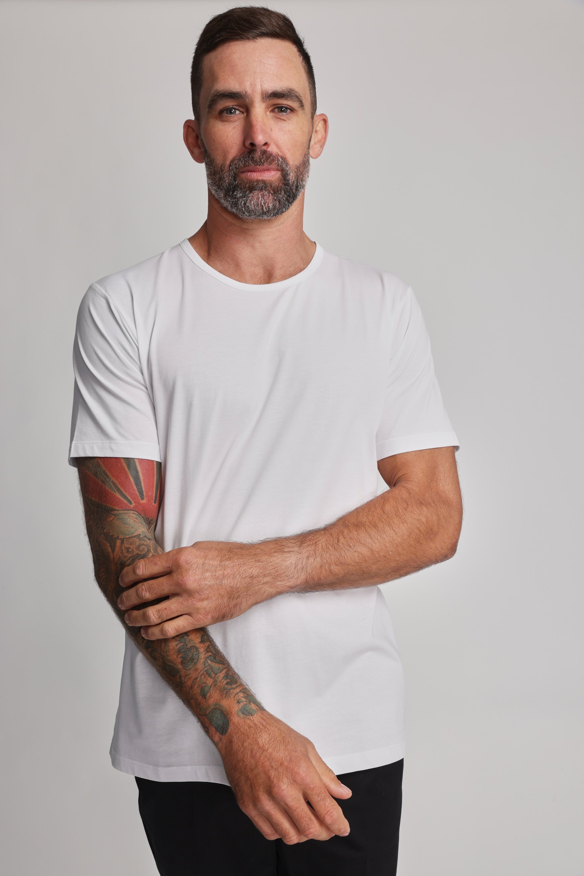 A man with short dark hair, a beard, and colorful left forearm tattoos stands against a plain background in a white Barkers Linton Supima Tee, looking directly at the camera with a neutral expression—a true wardrobe staple.