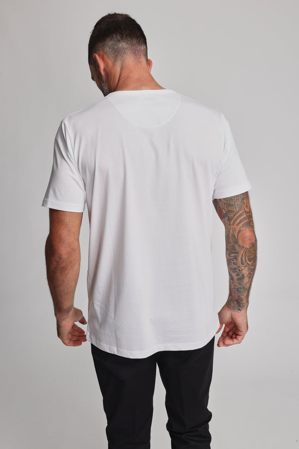 A man with short dark hair and a tattooed right arm stands with his back to the camera, wearing a plain white Barkers Linton Supima Tee and black pants against a light gray background.