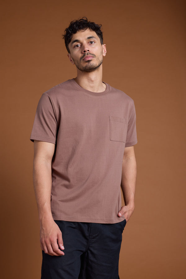 A man with short curly hair and light facial hair stands against a brown background, wearing a mauve Barkers Paddle Out Print Tee with a pocket and black pants, one hand in his pocket.