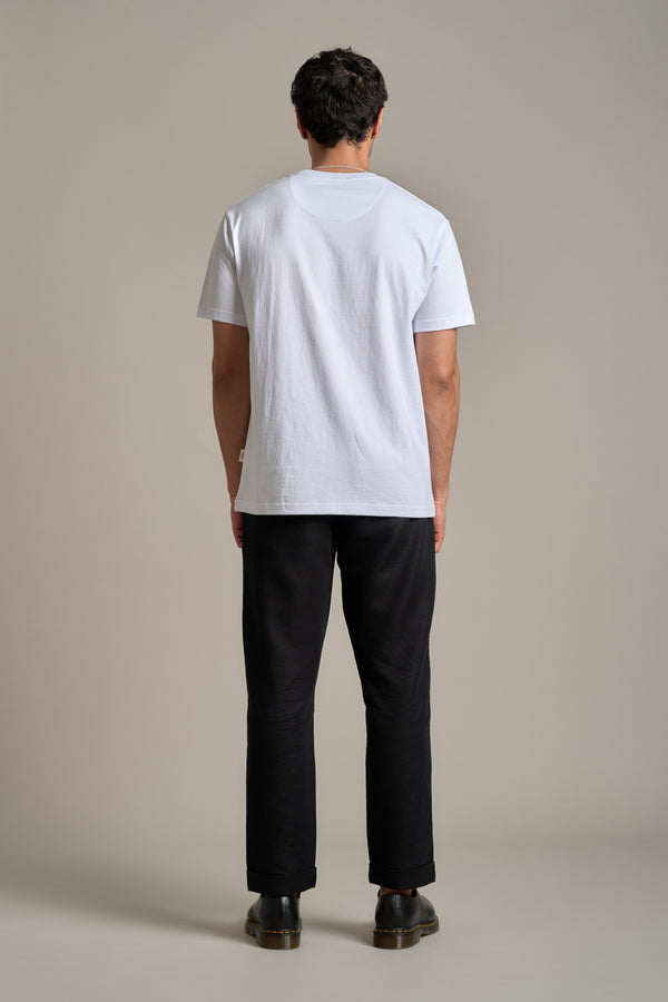 A person faces away from the camera, wearing the Barkers Boardriders Logo Tee in white, paired with black pants and shoes against a neutral background—an everyday essential look.