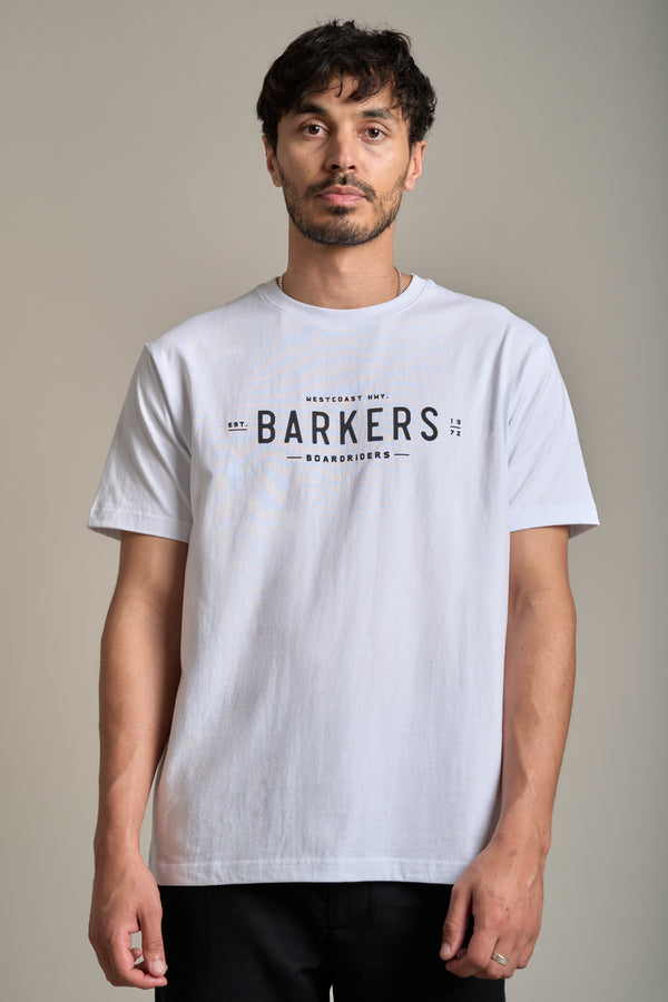 A man with short dark hair and facial hair models the Barkers Boardriders Logo Tee, an organic cotton essential featuring the bold Barkers logo. He stands facing forward against a neutral background to showcase the tee.