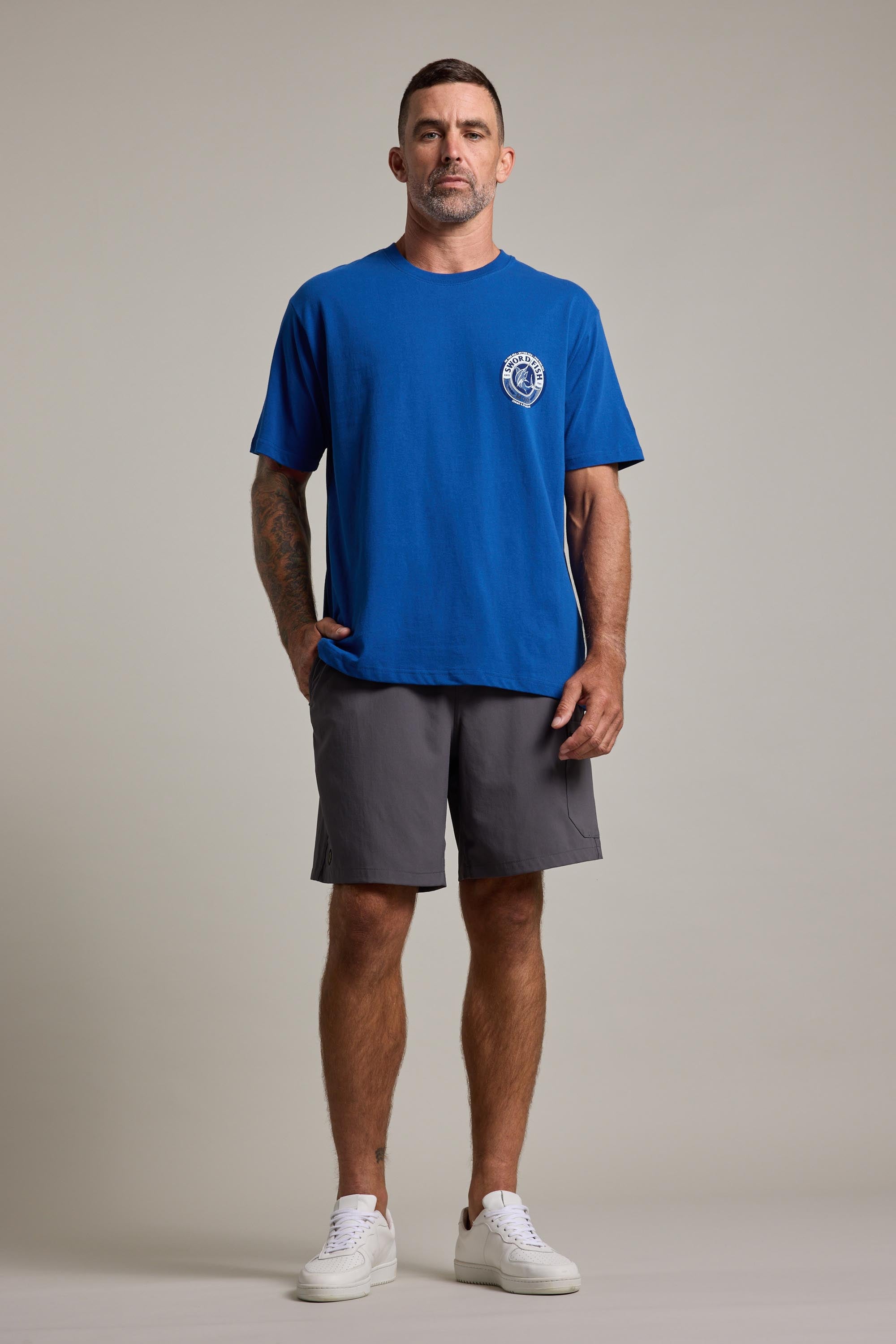 A man with short hair, a trimmed beard, and tattoos on his right arm stands against a neutral background wearing a blue Swordfish Mojo Logo Tee by Swordfish Mojo, featuring a small white crest on the chest, gray shorts, and white sneakers.