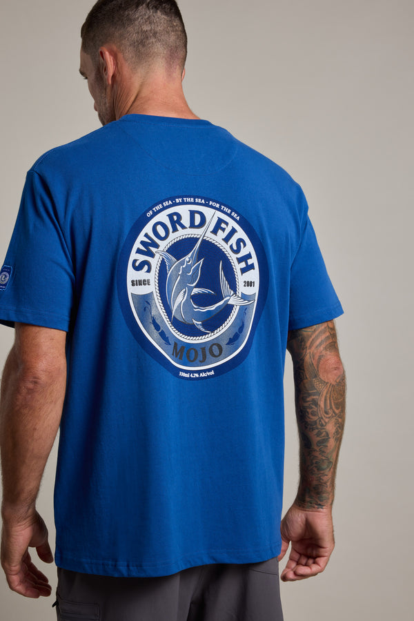 A man wears a blue Swordfish Mojo Logo Tee by Swordfish Mojo, featuring a large circular logo and fish graphic on the back. His tattooed right arm stands out against the plain background.