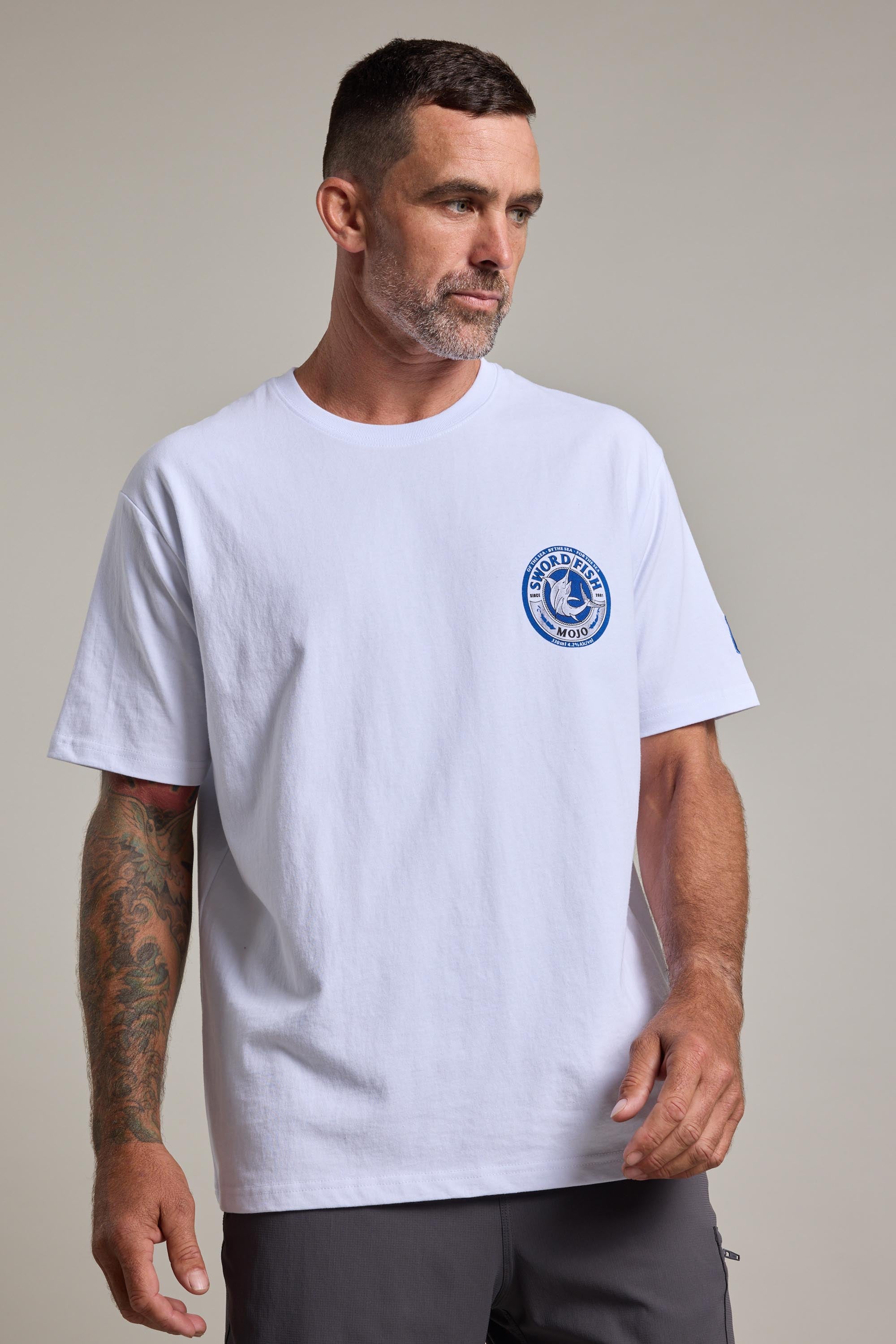 A man with short dark hair and a beard wears the Swordfish Mojo Logo Tee by Swordfish Mojo, featuring a small blue circular graphic on the left chest. He stands against a neutral background, his right arm covered in colorful tattoos.