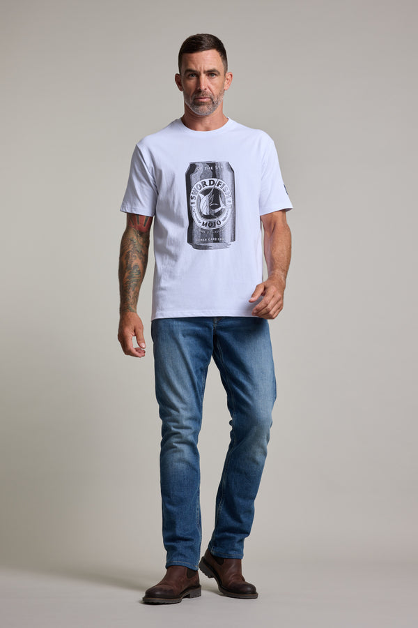 A man with short hair and right arm tattoos wears a Swordfish Mojo Beercan Tee by Swordfish Mojo, blue jeans, and brown boots, standing facing forward against a plain light gray background.