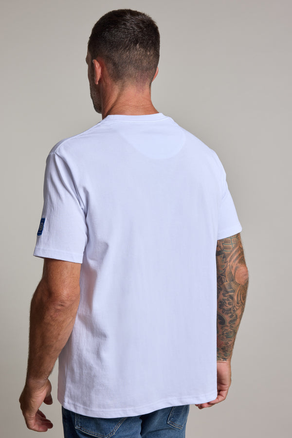 A man with short hair and visible arm tattoos stands with his back to the camera against a plain light gray background, wearing the Swordfish Mojo Beercan Tee by Swordfish Mojo and blue jeans.