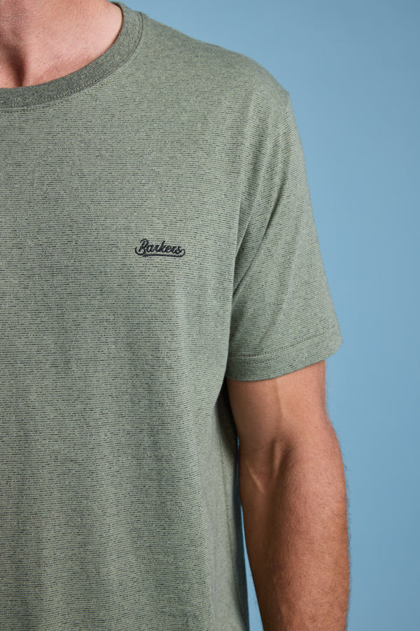 A person wearing the Barkers Bayside Emb Tee—an olive green, short-sleeved organic cotton T-shirt with "Barkers" embroidered on the left chest—stands against a solid light blue background.