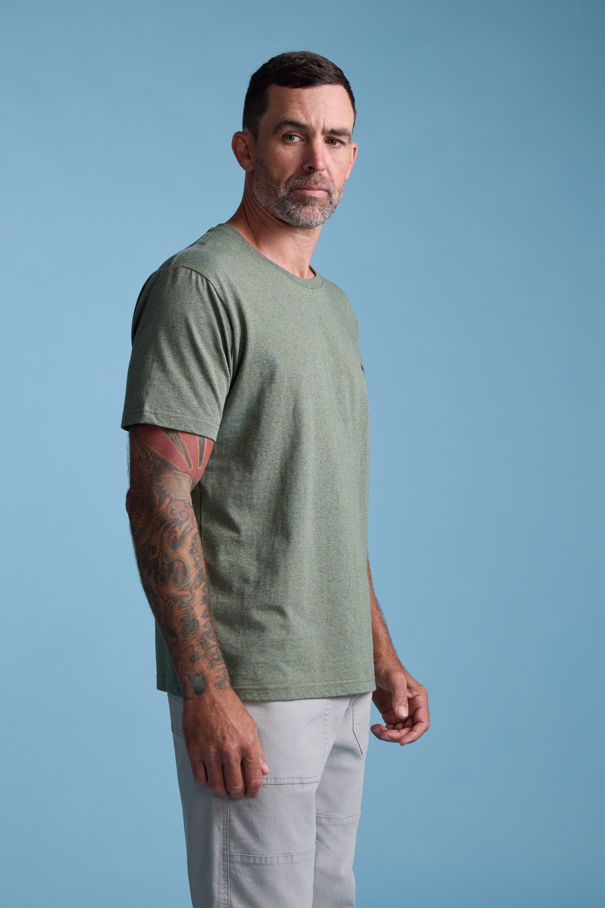 A man with short dark hair, a beard, and tattoos on his left arm stands against a blue background wearing the Barkers Bayside Emb Tee in light green and light gray pants, looking slightly to the side.