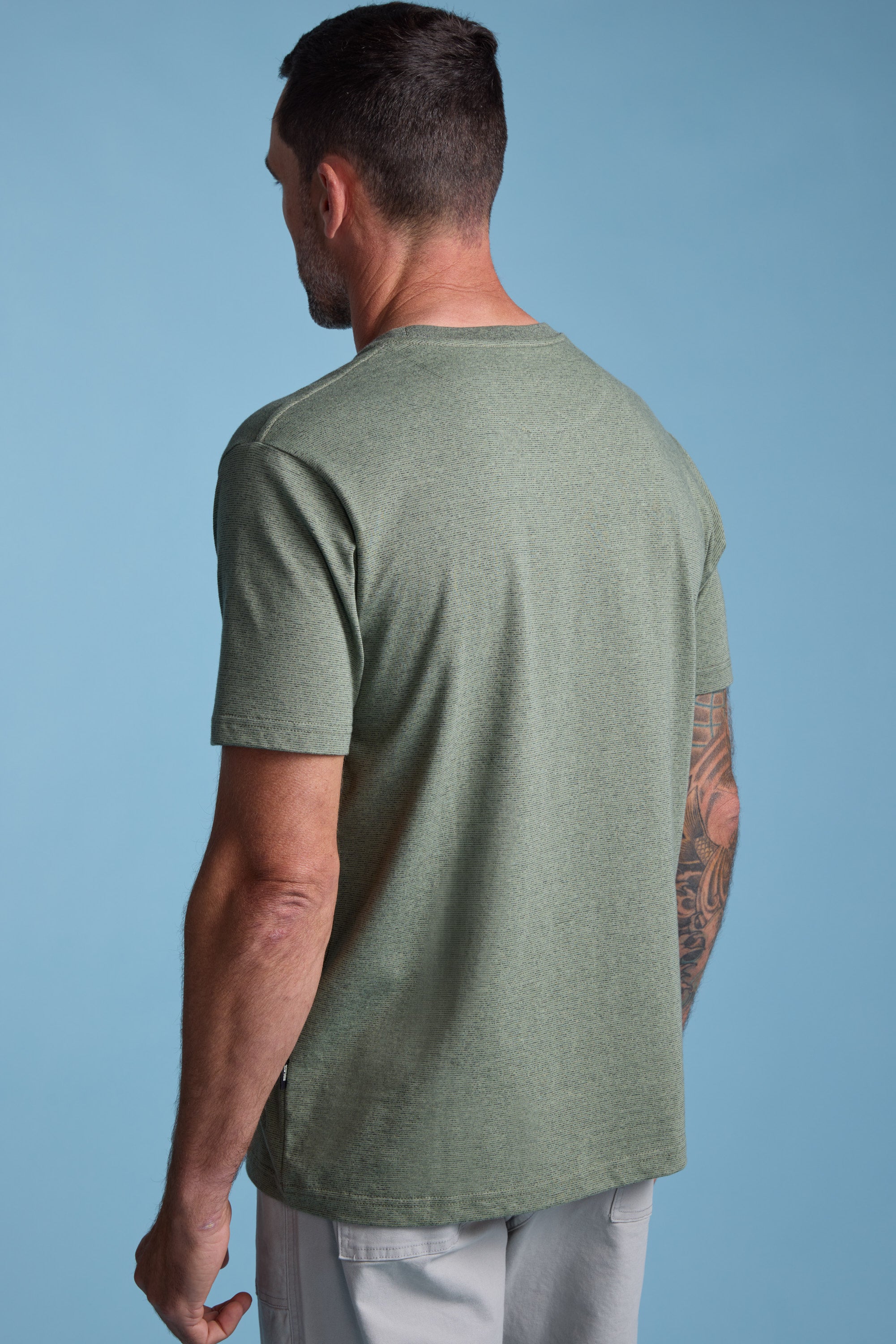 A man with short dark hair, seen from behind, wears the Barkers Bayside Emb Tee in light green and light grey shorts against a plain blue background. One of his arms features a visible tattoo.