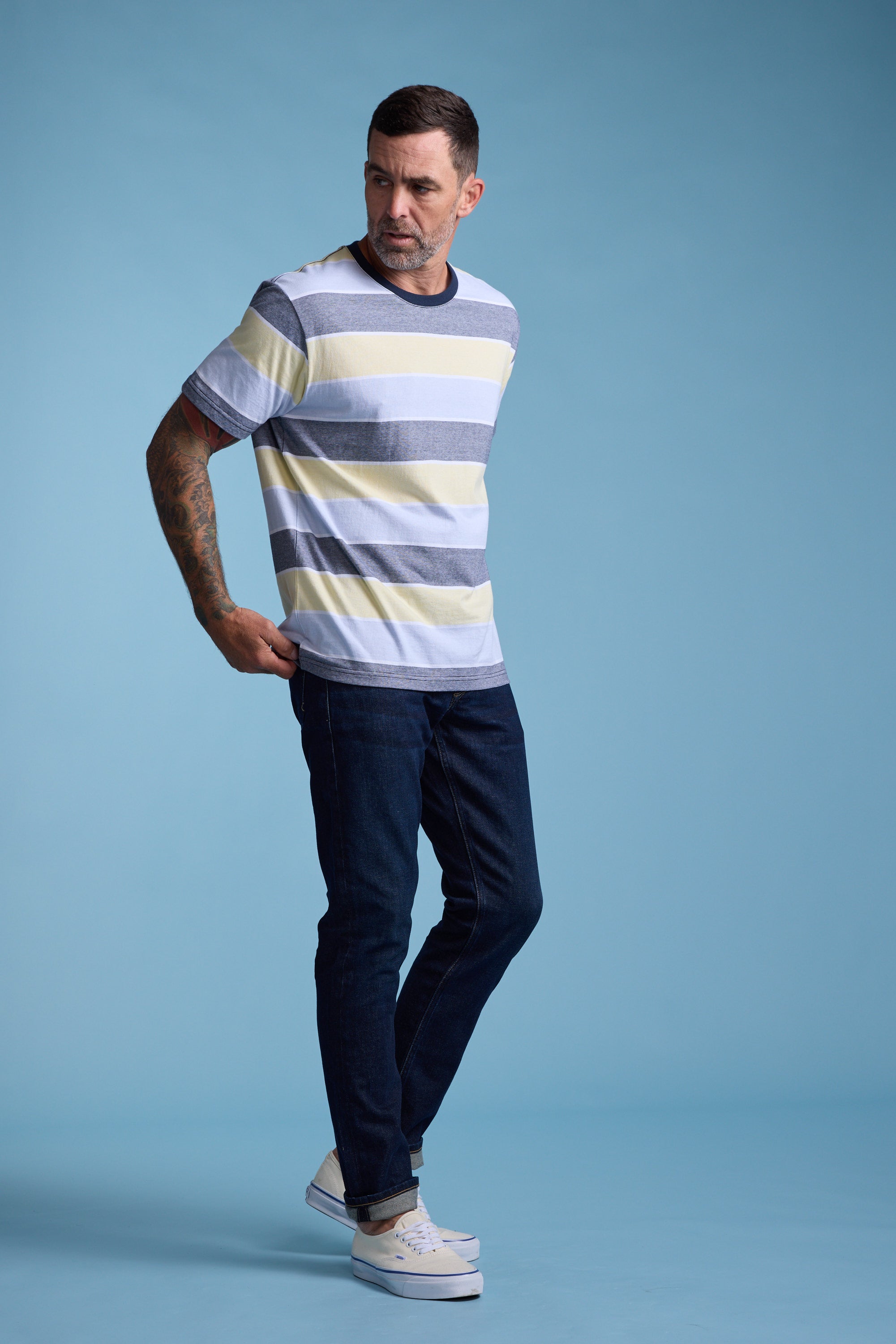 A man wearing the Barkers Asbrooke Stripe Tee, paired with dark jeans and white sneakers, stands against a blue background, looking left with one hand behind his back.