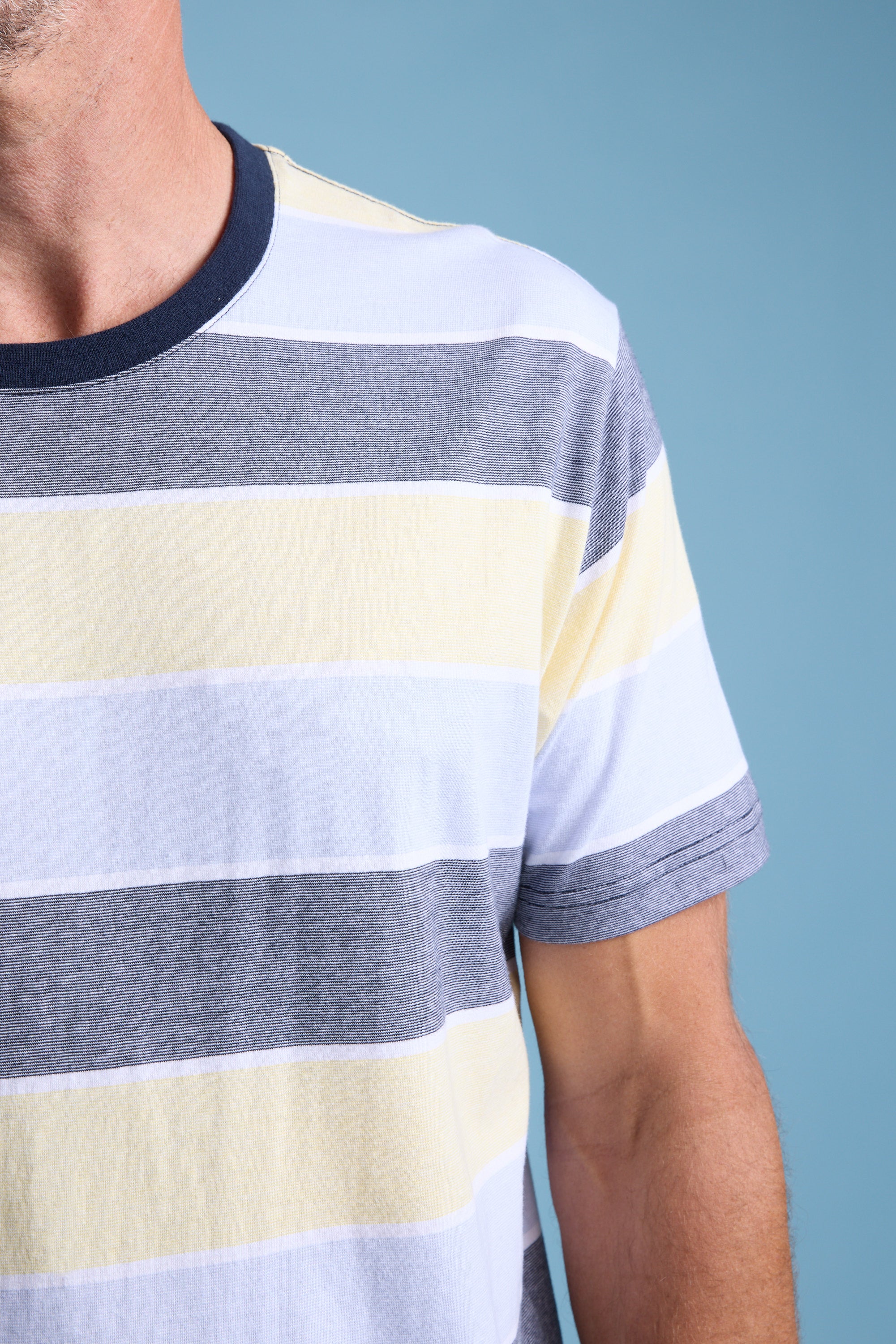 A close-up of a person wearing the Barkers Asbrooke Stripe Tee in gray, white, and pale yellow against a blue background. Only the upper torso and part of one arm are visible, highlighting the tee’s relaxed fit and style.