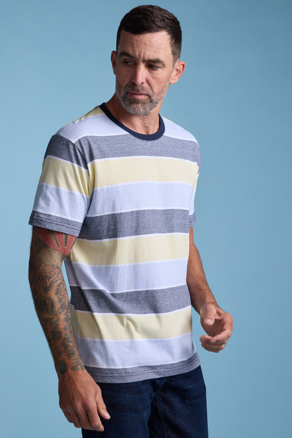 A man with short dark hair and a trimmed beard wears the Barkers Asbrooke Stripe Tee in yellow, white, and gray, revealing his tattooed right arm as he stands against a plain blue background.