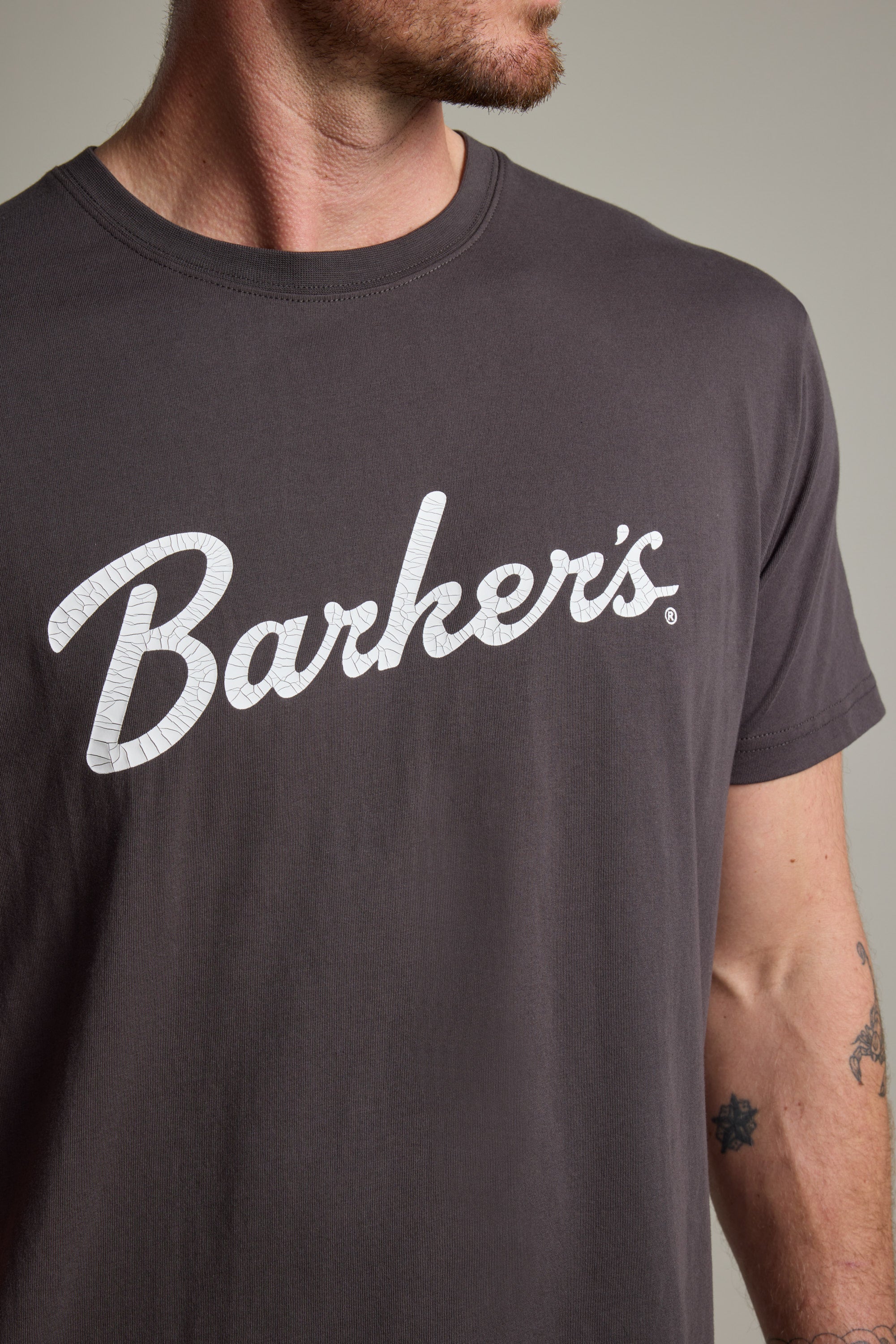 A person wears the Barkers Finely Tuned Print Tee, an organic cotton t-shirt in dark gray with a vintage crackle "Barkers" logo. The photo shows the upper torso and part of a tattooed arm.