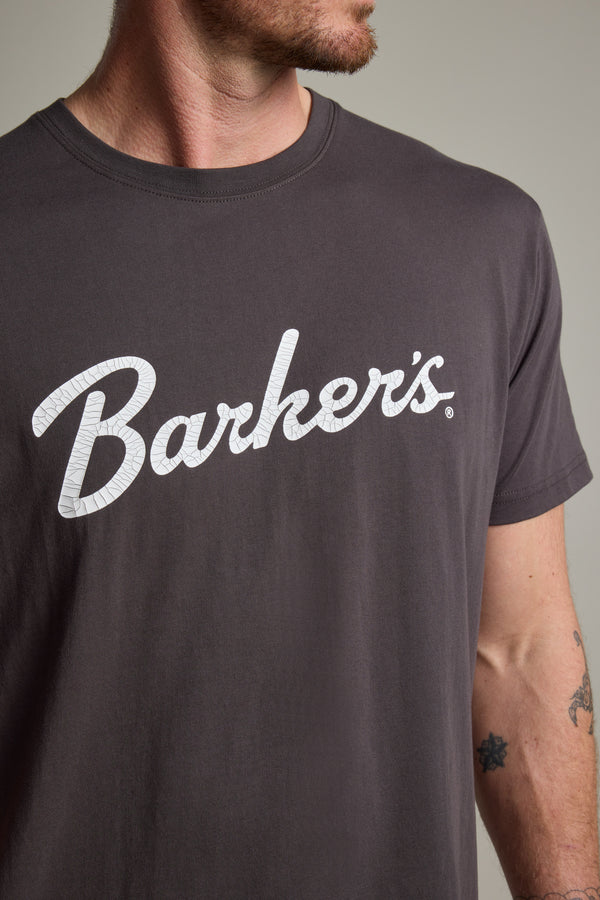 A person wears the Barkers Finely Tuned Print Tee, an organic cotton t-shirt in dark gray with a vintage crackle "Barkers" logo. The photo shows the upper torso and part of a tattooed arm.