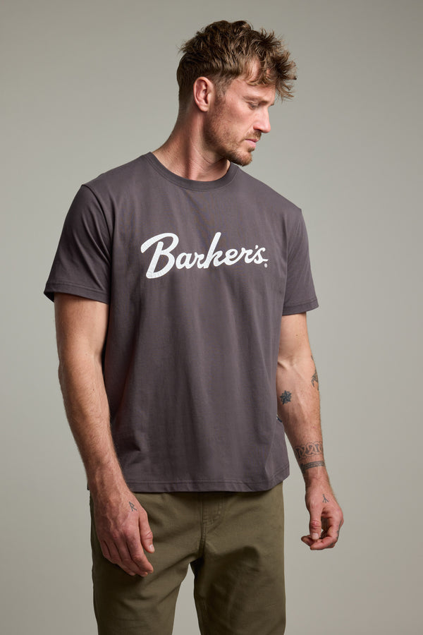 A man with short, tousled hair and visible arm tattoos wears a gray Barkers Finely Tuned Print Tee in organic cotton and olive pants, standing against a plain light background while looking to his left.