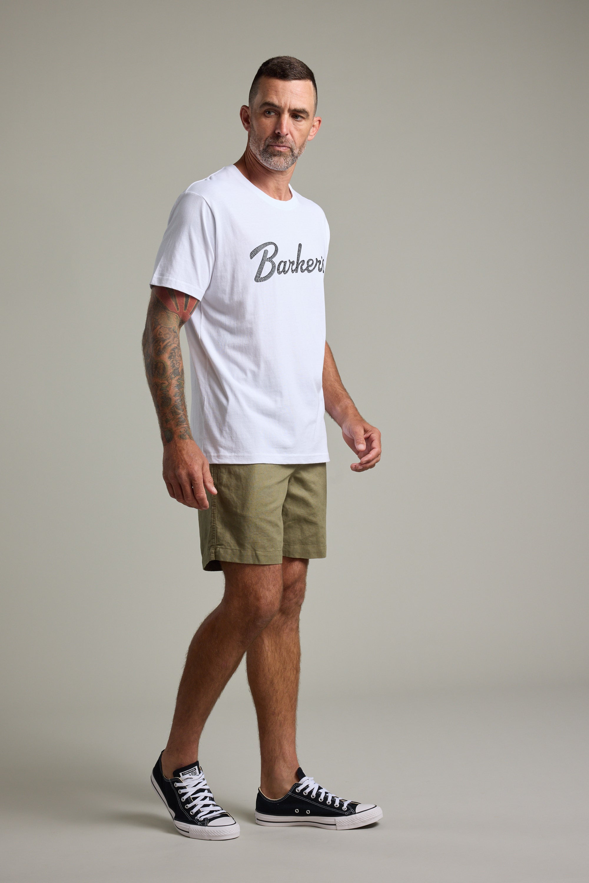 A man with short hair and a beard poses in a studio, wearing the Barkers Finely Tuned Print Tee in white, khaki shorts, and black sneakers. He displays tattoos on his arms while standing against a plain light background.