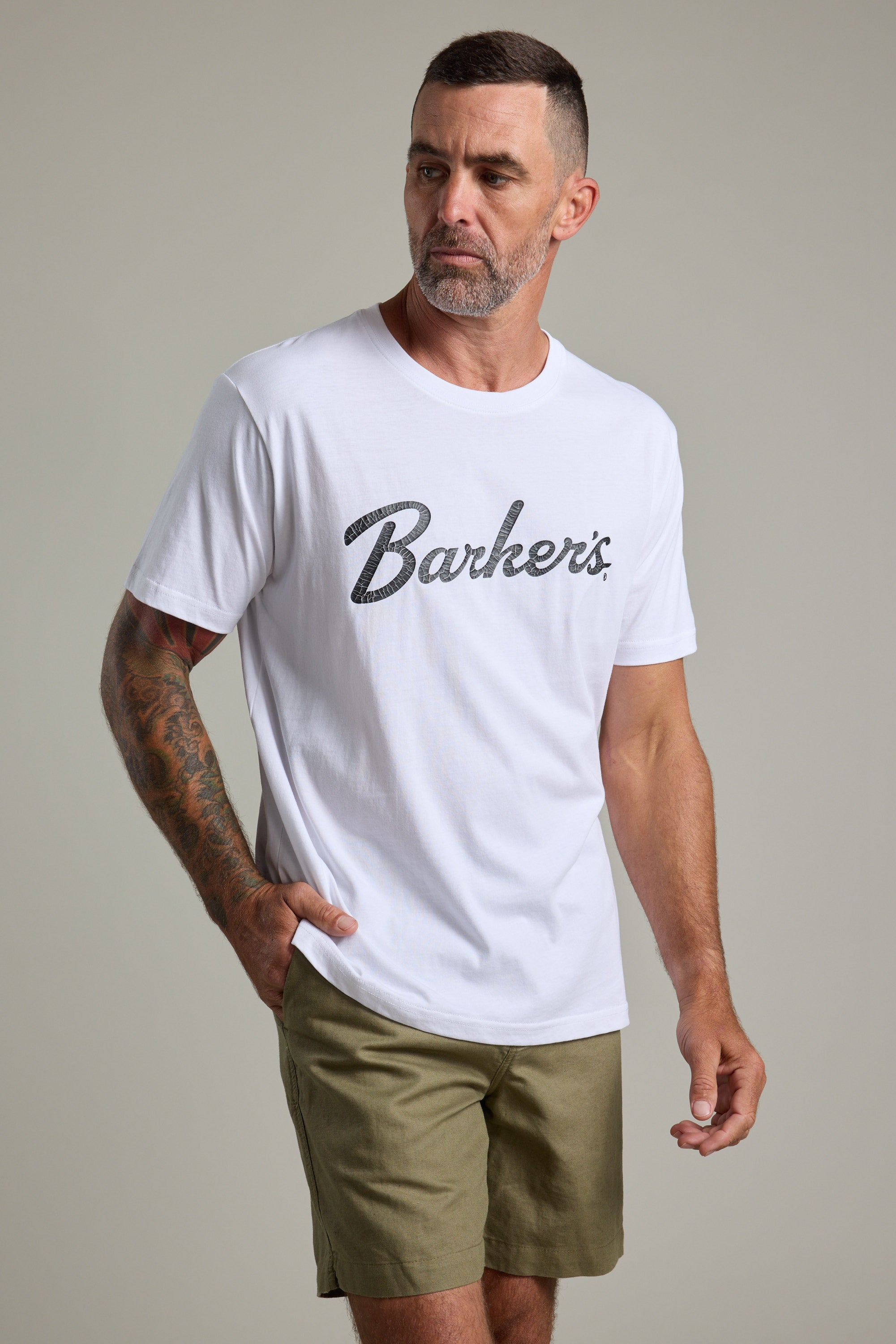 A man with short hair, a beard, and a tattooed left arm wears the Barkers Finely Tuned Print Tee in white with khaki shorts. He stands against a plain light background, looking slightly to his left.