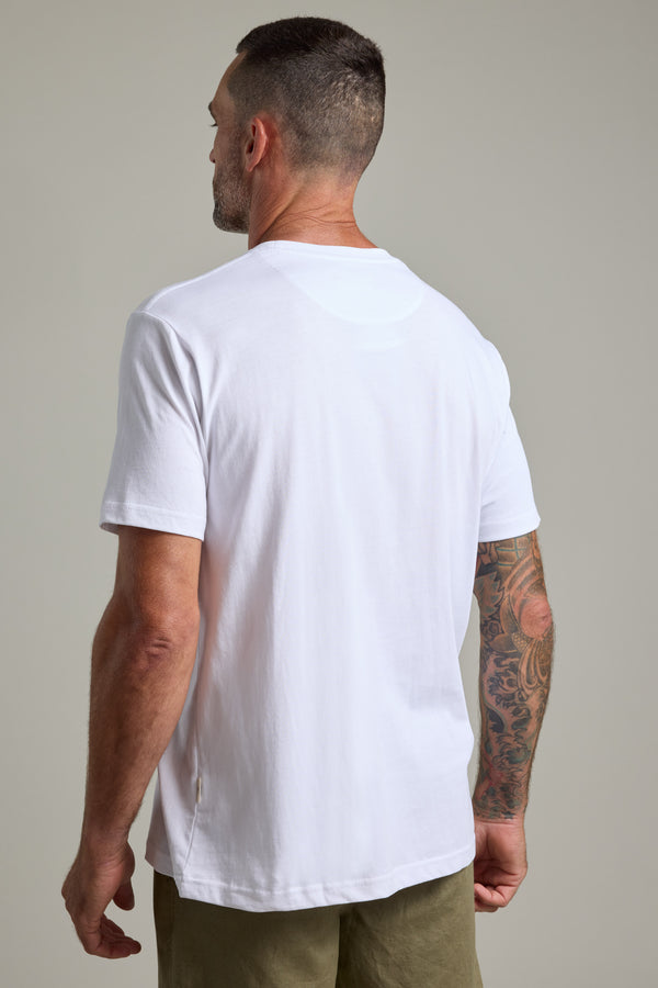 A man with short hair and tattoos on his right arm faces away, displaying the back of his Barkers Finely Tuned Print Tee in white, styled with olive green shorts against a plain background.