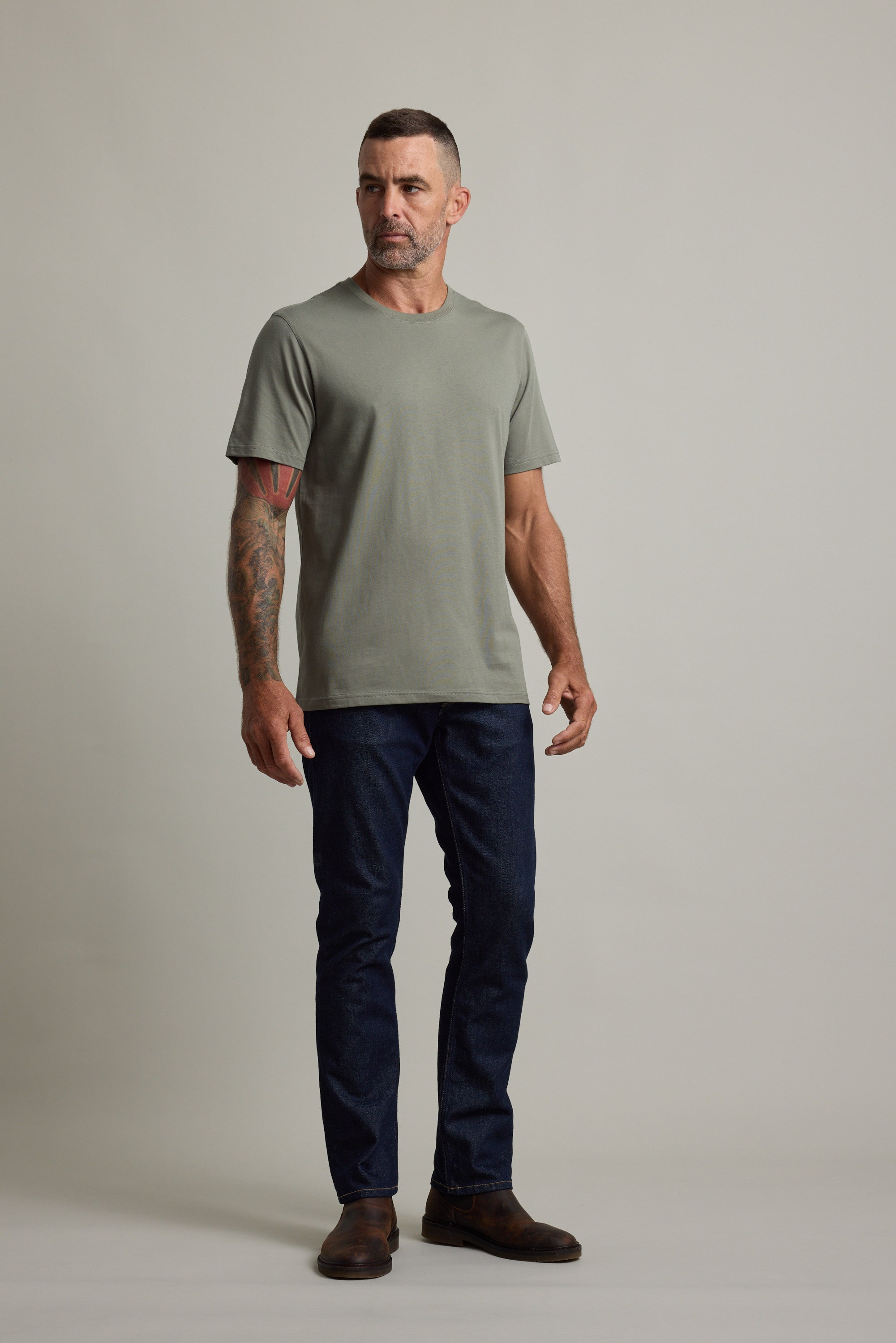 Tattooed man wears Barkers Good Earth Cotton Essential Tee, dark jeans, and brown boots.