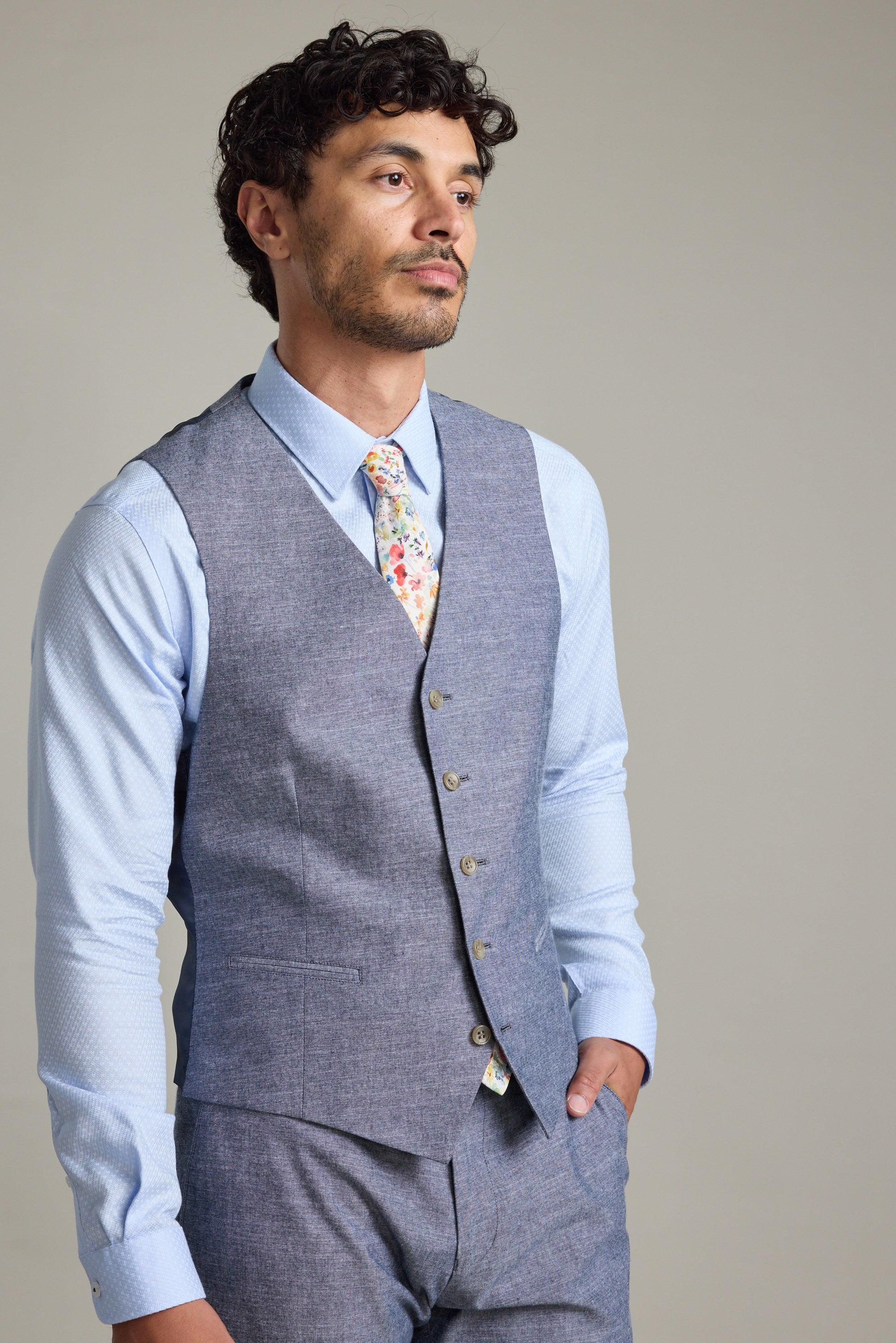A man stands with one hand in his pocket, wearing Barkers' Brighton Cotton Waistcoat with an adjustable back buckle, a light blue dress shirt, matching gray pants, and a floral tie, gazing slightly to the side against a plain background.