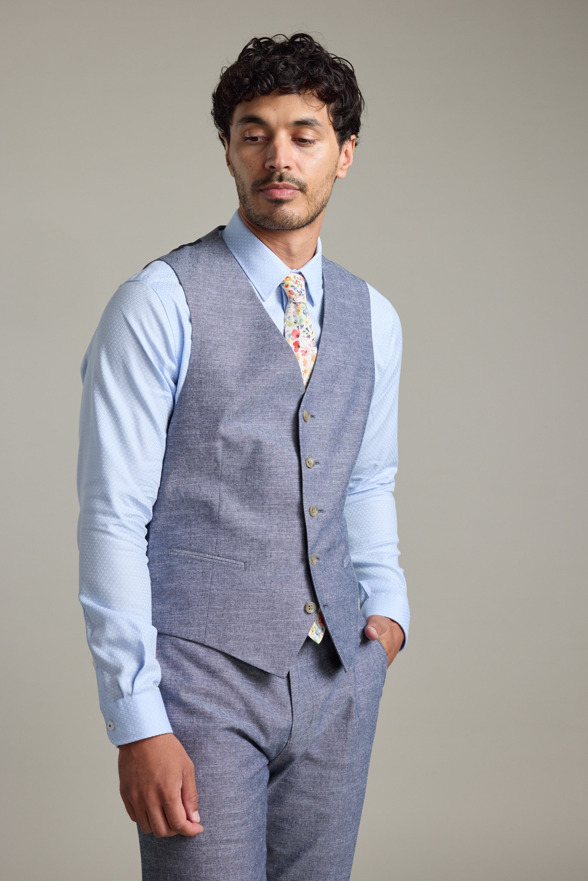 A man with short, curly hair wears the Barkers Brighton Cotton Waistcoat in gray over a light blue shirt, with matching slacks and a floral tie. He stands with one hand in his pocket, glancing downward and to the side.