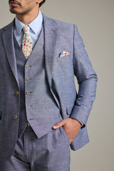 A man wears the Barkers Brighton Cotton Waistcoat in light blue as part of a three-piece suit with a floral tie, posing with one hand in his pocket. The waistcoat includes an adjustable back buckle for a perfect fit.
