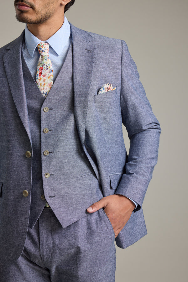A man wears the Barkers Brighton Cotton Waistcoat in light blue as part of a three-piece suit with a floral tie, posing with one hand in his pocket. The waistcoat includes an adjustable back buckle for a perfect fit.