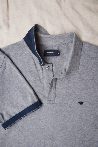 The Barkers Regent Pique Polo is a gray men's polo crafted from soft organic cotton, featuring a navy collar and sleeve trim, buttoned placket, and a small chest logo. It’s displayed flat on a light fabric surface.