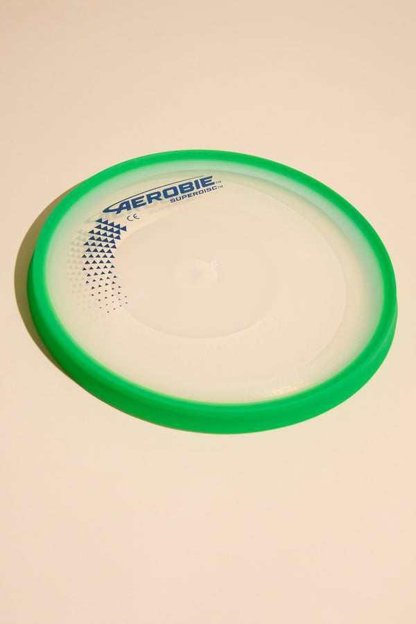An Aerobie Superdisc by Other, featuring a round shape, translucent center, and bright green outer ring, lies flat on a light beige surface.