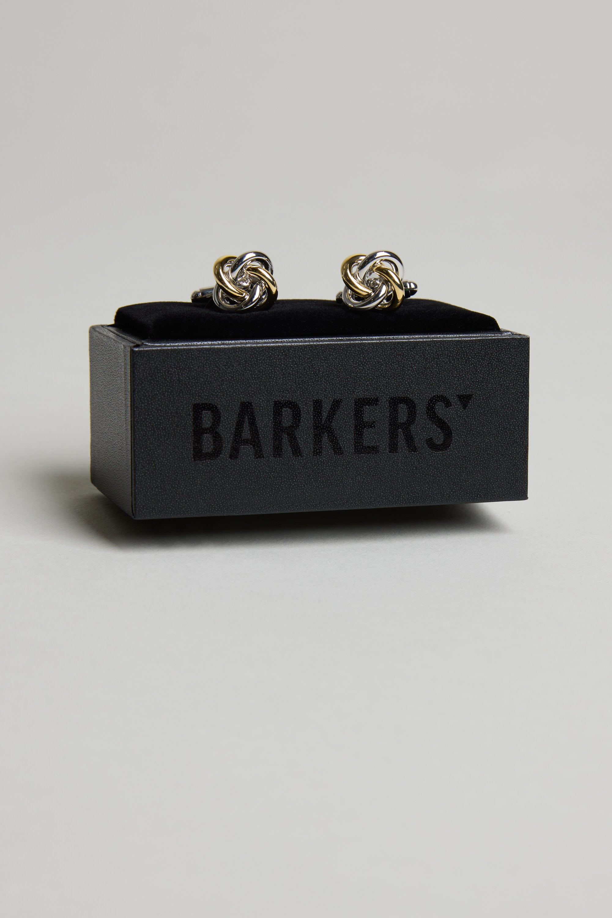 A pair of Barkers Knot Cufflink in silver and gold, elegantly displayed on a black velvet stand inside a sleek box with BARKERS branding. The perfect formal accessory for any special occasion.