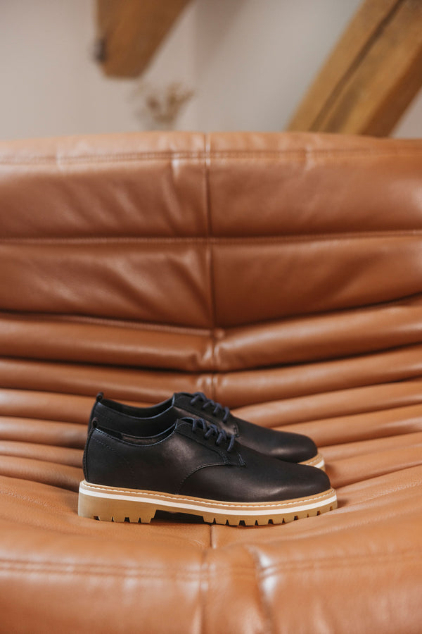 A pair of CLAE Logan shoes in premium leather with tan soles on a brown leather couch.
