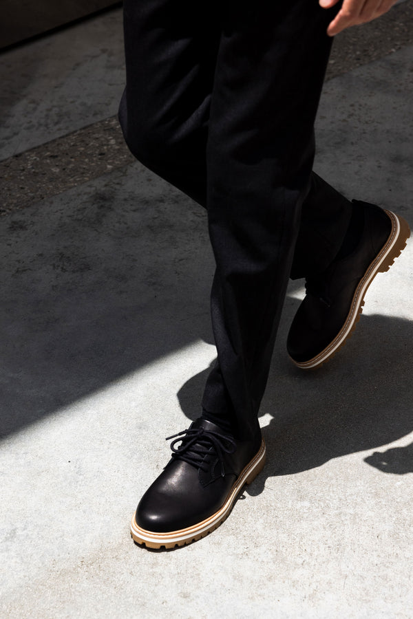 Person wearing Clae Logan premium leather black dress shoes and black pants on concrete.