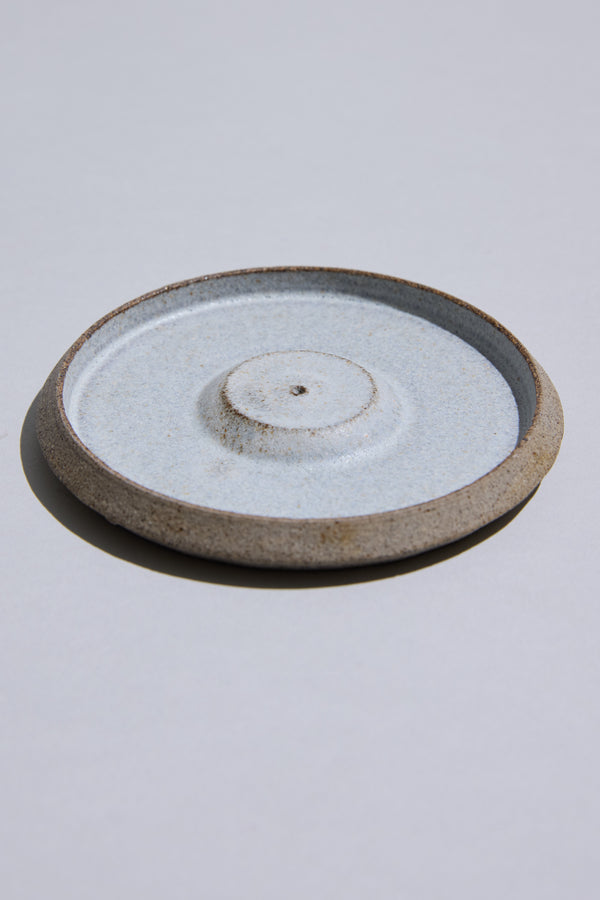 The Studio Foon Incense Dish by Other is a round, shallow New Zealand ceramic plate with a raised center and unglazed rim, featuring a light speckled glaze made from natural clay. It rests on a plain light gray surface.