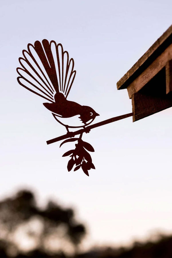 The Other Piwakawaka / Fantail features a Corten steel bird perched on a branch beside a wooden structure, blending with the clear sky and blurred trees behind.