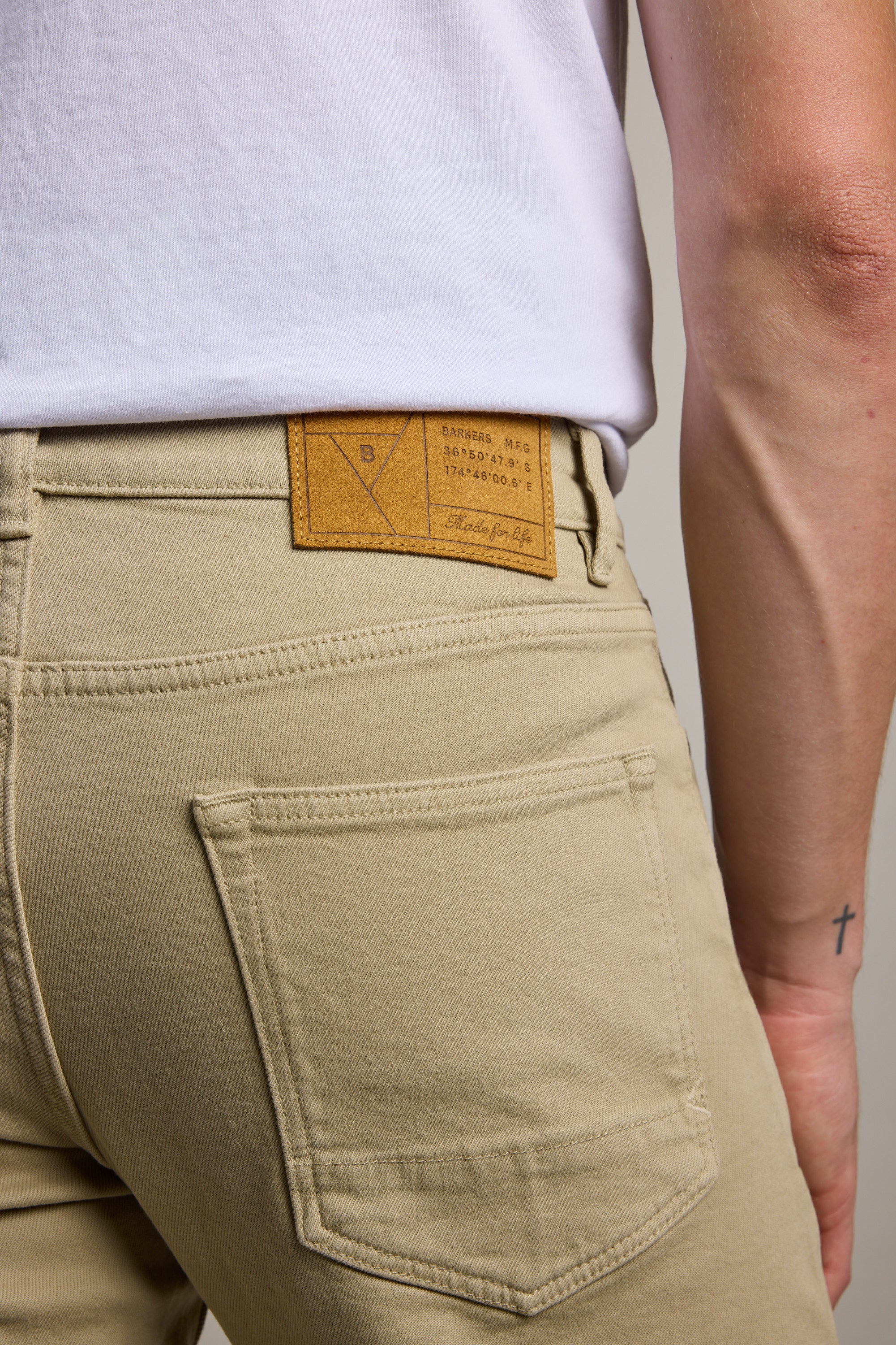 A person is shown from the waist down wearing Barkers Rambler Slim Coloured Jeans in beige with a brown leather patch, visible back pocket, and a white shirt; a small cross tattoo appears on their left wrist.