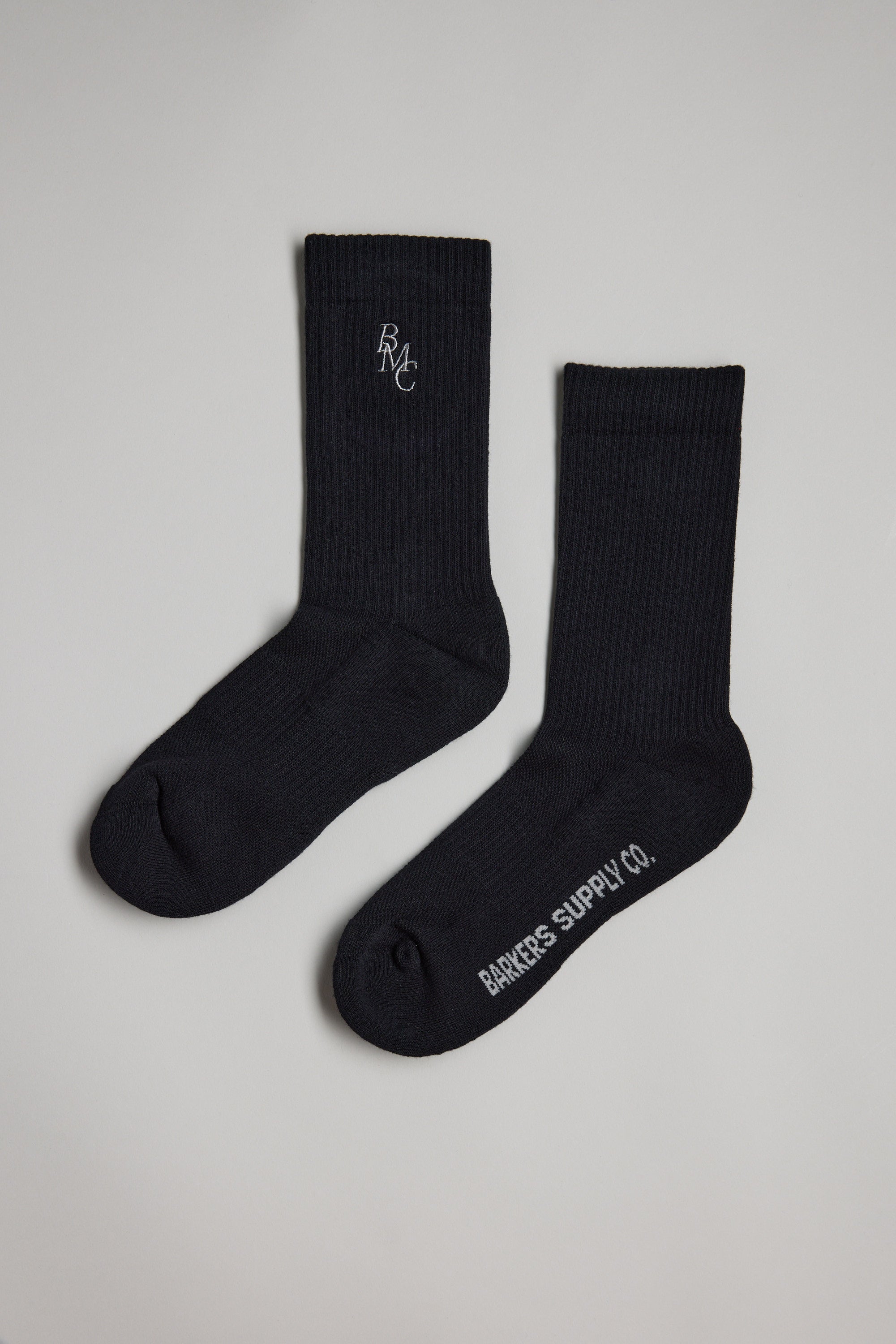 Two black Barkers BMC Ribbed Socks are laid flat on a light gray surface—one features "BMC" embroidered near the top, and the other has "Barkers Supply Co." printed in white on the sole.