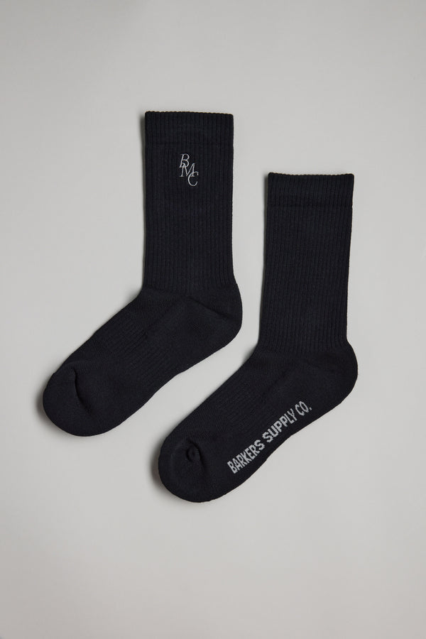 Two black Barkers BMC Ribbed Socks are laid flat on a light gray surface—one features "BMC" embroidered near the top, and the other has "Barkers Supply Co." printed in white on the sole.