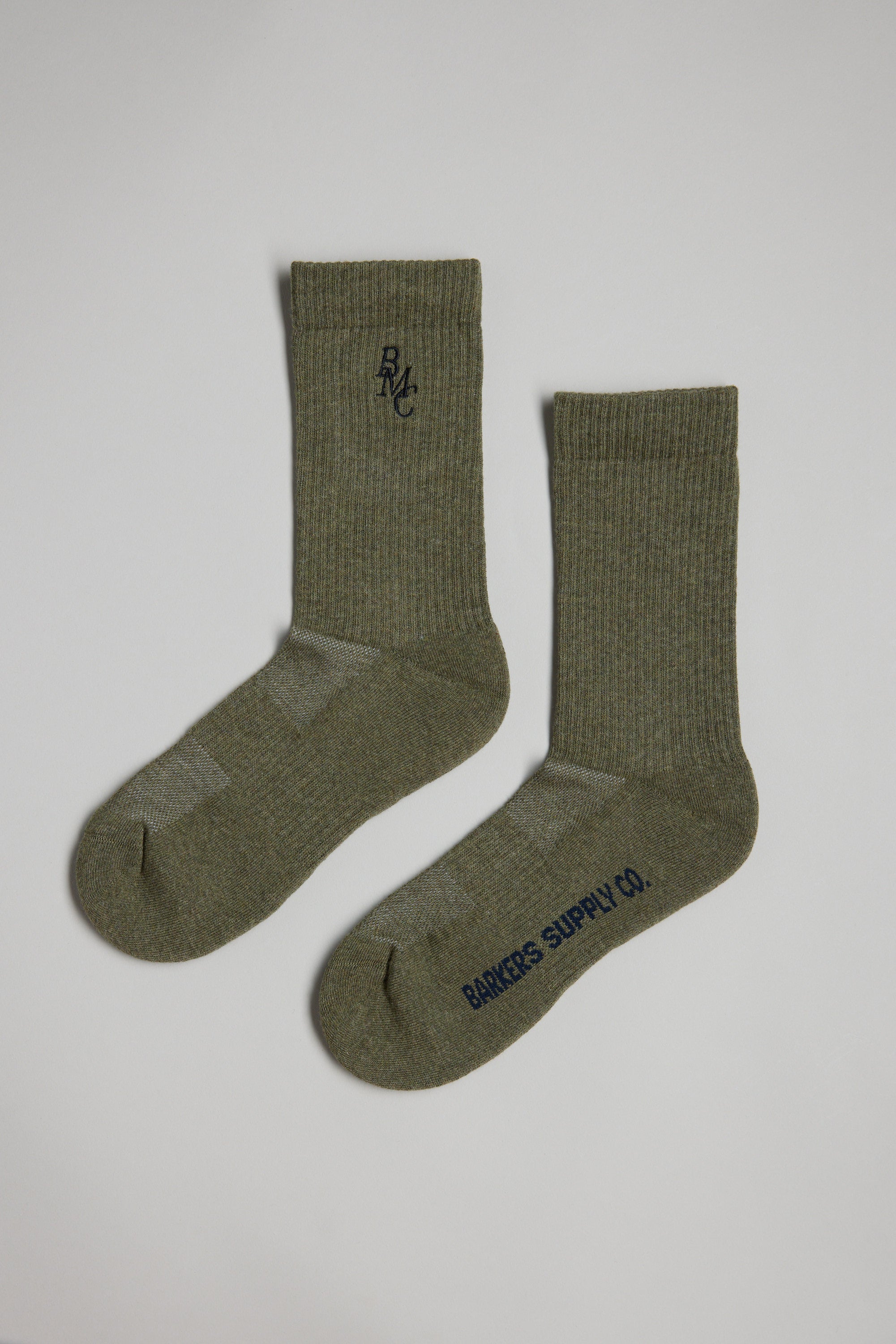 A pair of olive green BMC Ribbed Socks by Barkers, featuring "RMC" embroidery near the top of one sock and "BANKERS SUPPLY CO." in black on the sole of the other, laid flat on a light background.